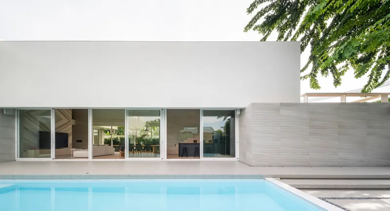 White facade with sliding glass doors opening to a pool terrace under overhanging tree branches