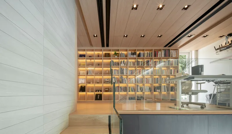 Interior view of backlit bookshelf wall with timber ceiling and glass balustrade overlooking kitchen