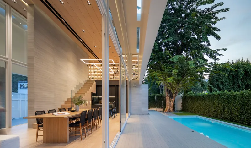 Interior view through floor-to-ceiling glass doors showing timber-lined dining space and pool deck at dusk