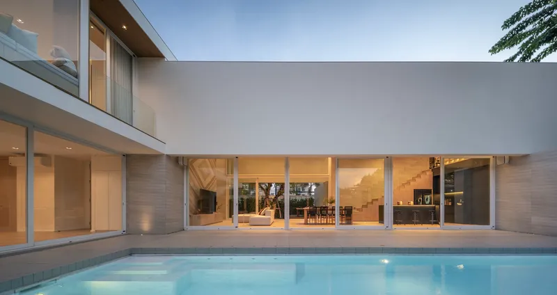 Pool terrace surrounded by illuminated glass-walled interior spaces under an overcast evening sky