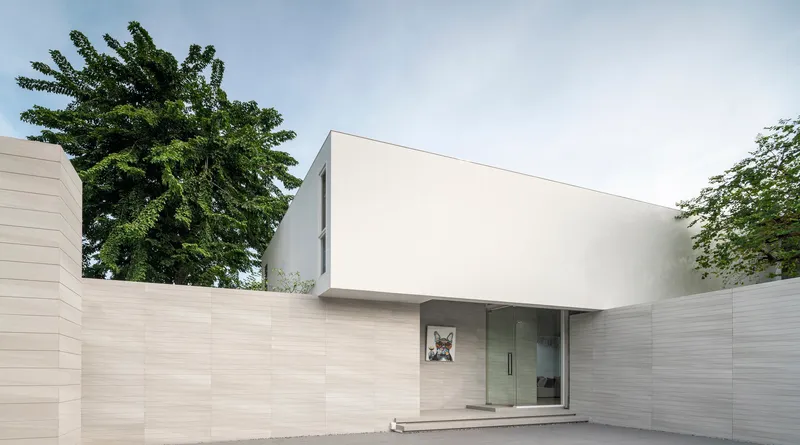 Street view of the white upper volume floating above horizontal stone walls and mature trees