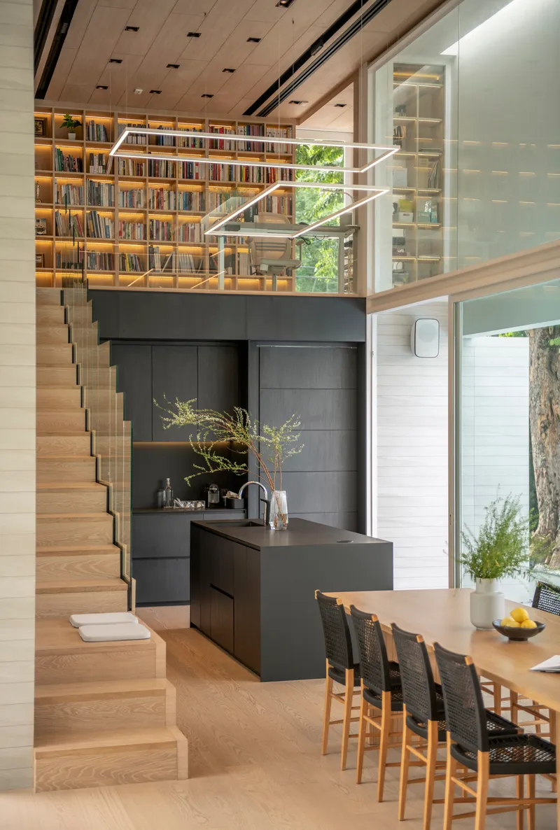 Double-height interior showing timber staircase ascending to mezzanine library above black kitchen island
