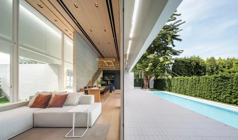 Open-plan living area with timber ceiling and outdoor pool terrace framed by mature trees and hedge