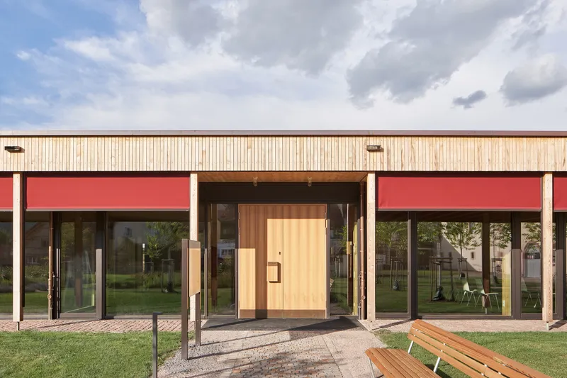 Entrance facade with red fabric awnings above glazed openings and vertical timber cladding under clouds