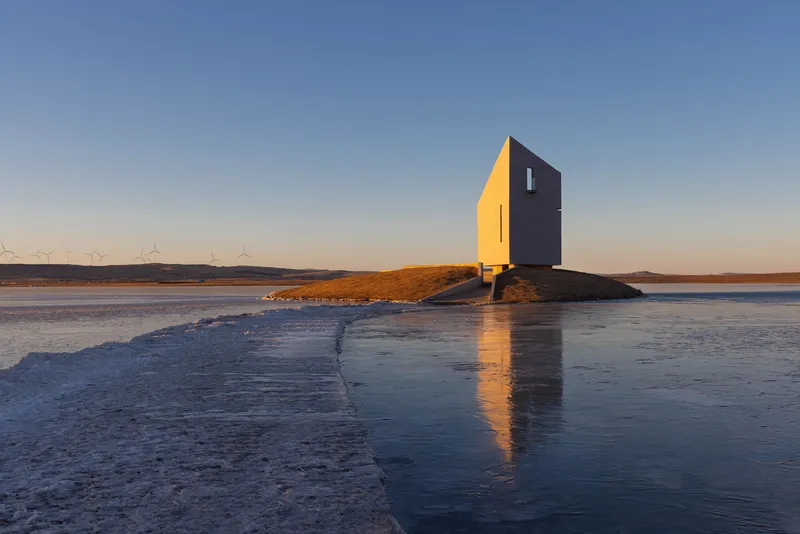 The two-tone facade reflected in the icy water with wind turbines on the horizon