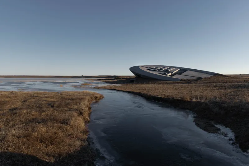 The elliptical roof structure rising from grassland beside a meandering frozen stream under clear sky