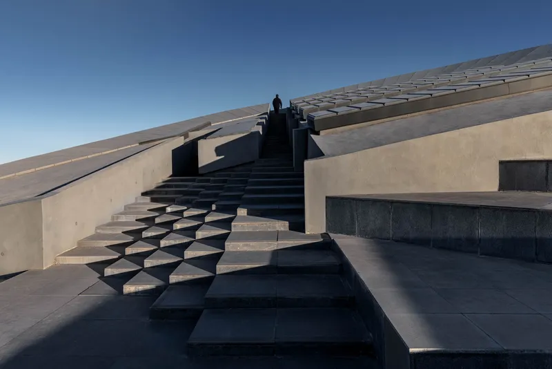 Cantilevered concrete staircase with geometric treads ascending the sloped roof surface with a lone figure