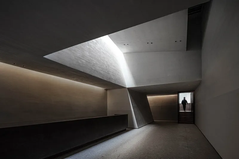 Interior gallery space with angular concrete walls and a silhouetted figure ascending the distant stair
