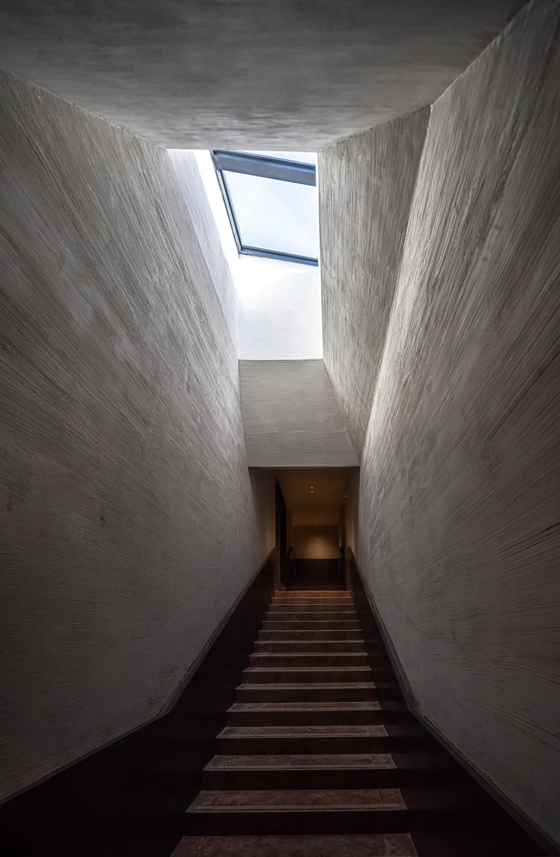 Timber staircase rising through a tall concrete light well with angled skylight above