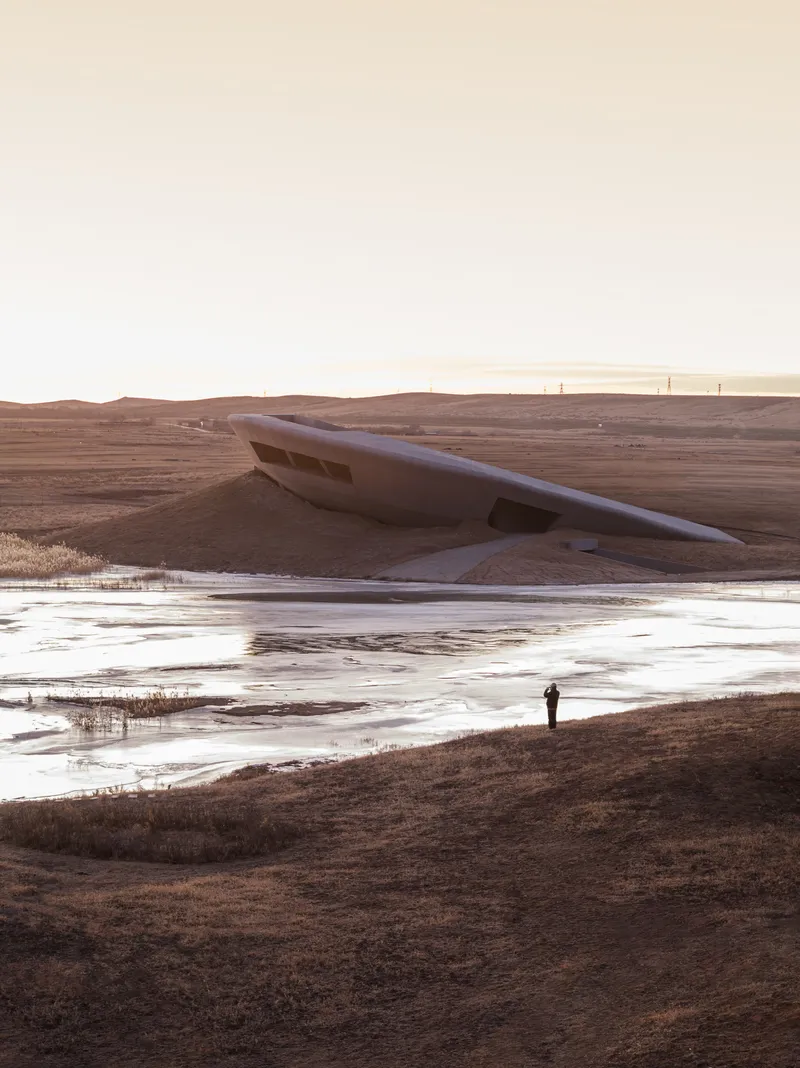 The fluid concrete roof form rising from the frozen shoreline with a lone visitor at sunset