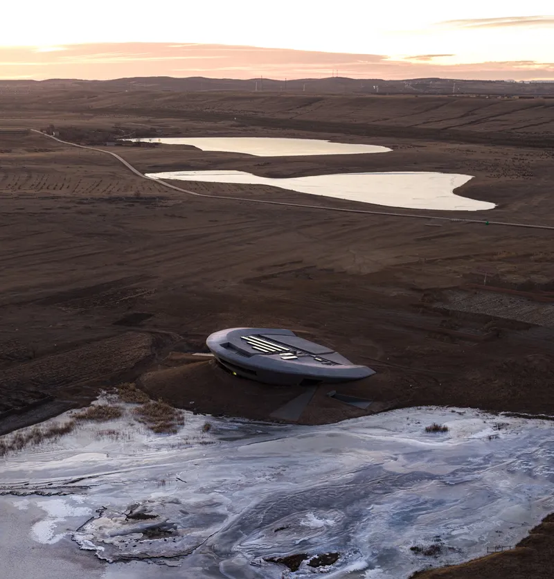Drone view of the sculptural roof form set in the brown steppe between frozen ponds at sunset