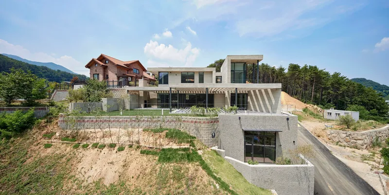 Terraced concrete house stepping down a hillside with retaining walls and adjacent red-roofed buildings