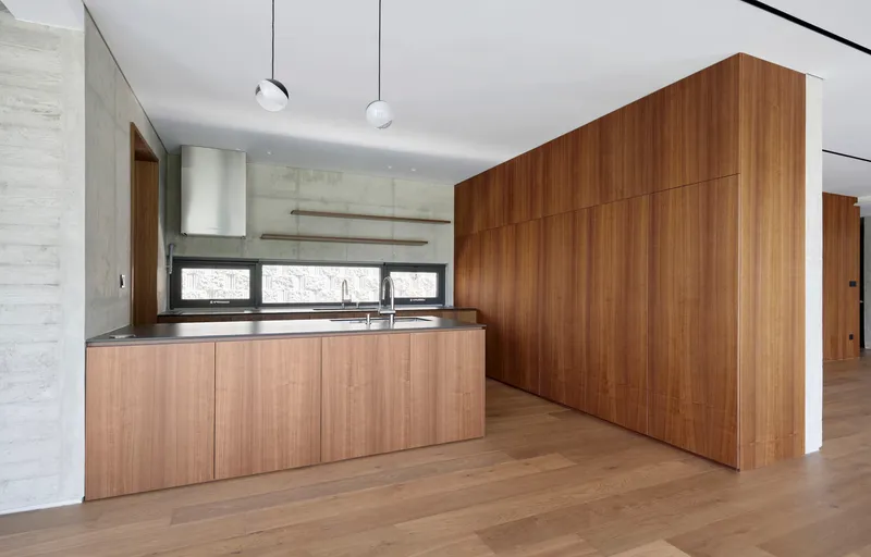 Kitchen with wood veneer island and wall cabinetry beside board-formed concrete walls and globe pendant lights