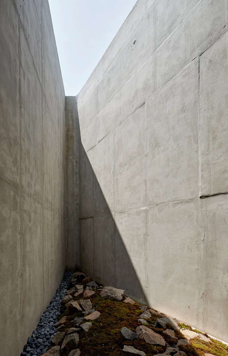 Narrow exterior courtyard between board-formed concrete walls with rocks and moss on the ground