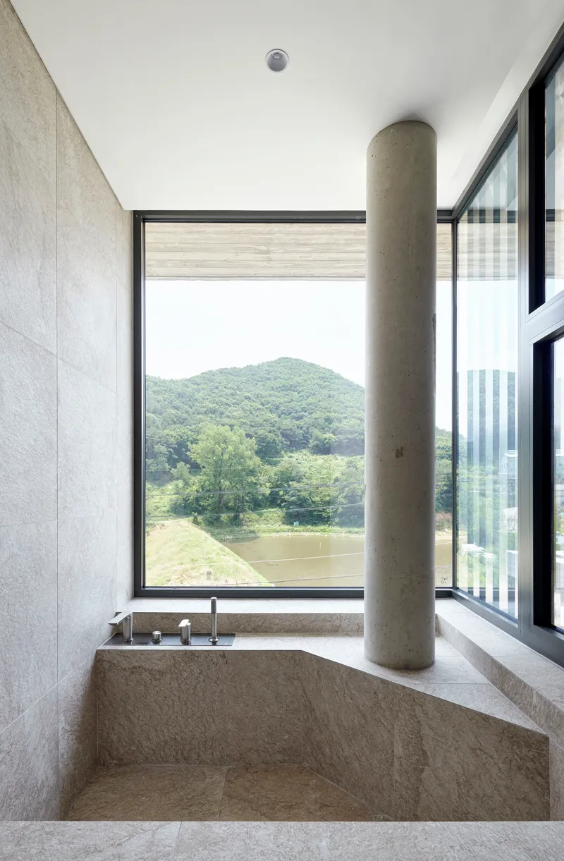 Stone bathtub with cylindrical concrete column beside a corner window framing a tree and distant mountain