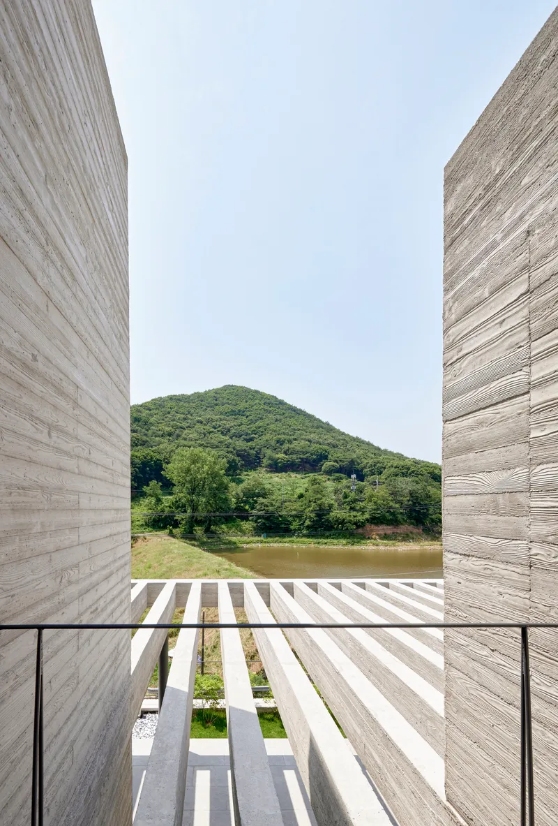 View through board-formed concrete walls toward a slatted timber deck, pond, and green hills beyond