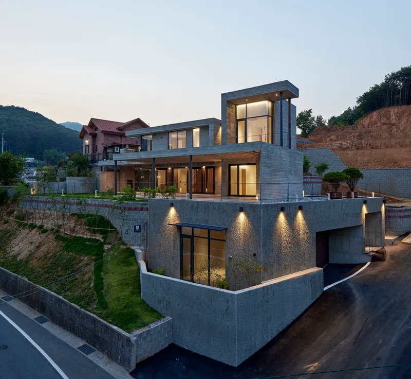 Three-level concrete residence on sloped site with glazed openings illuminated at dusk