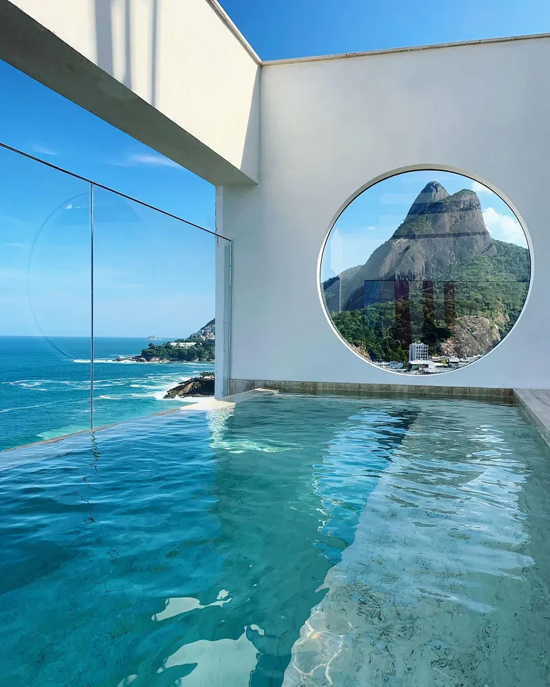 Infinity pool with circular porthole window framing a coastal mountain beyond the blue water