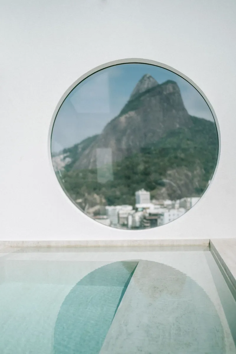 Round window overlooking a forested mountain peak with poolside reflection below