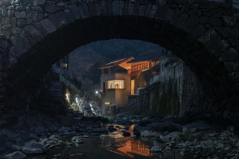 View through a stone arch bridge to the timber-clad facade illuminated at dusk above a rocky stream