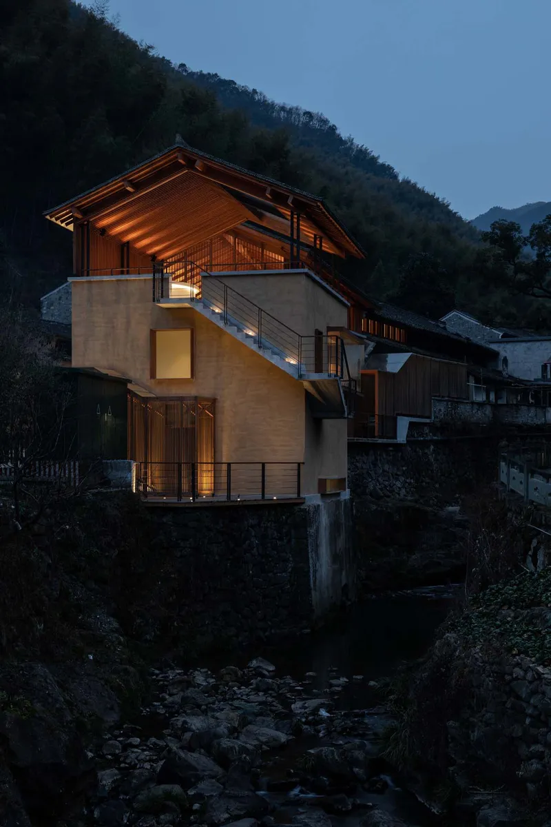 Three-story residence with rammed earth walls and timber roof illuminated above a rocky stream at dusk