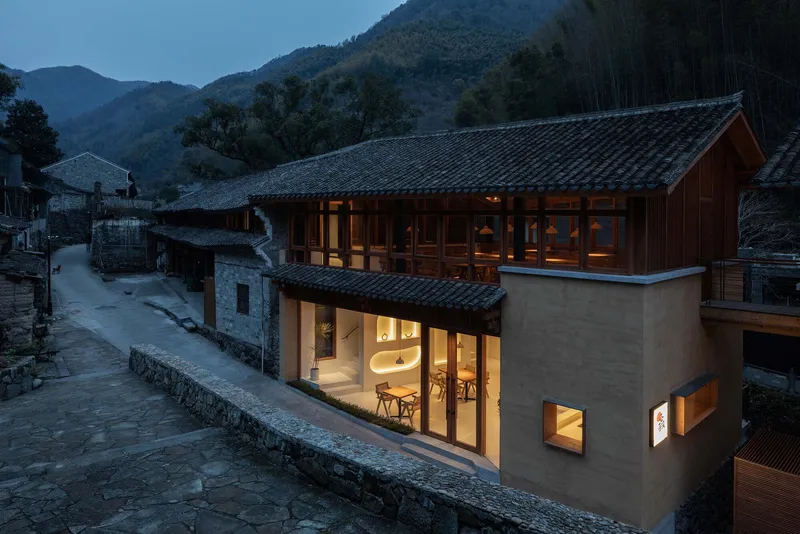 Curved facade with large glazed openings and tile roof alongside a stone-paved village path at twilight
