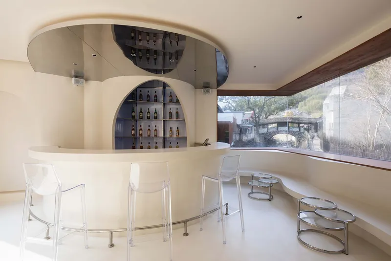 Curved bar with arched display niche and transparent acrylic chairs overlooking a hillside through glazed window
