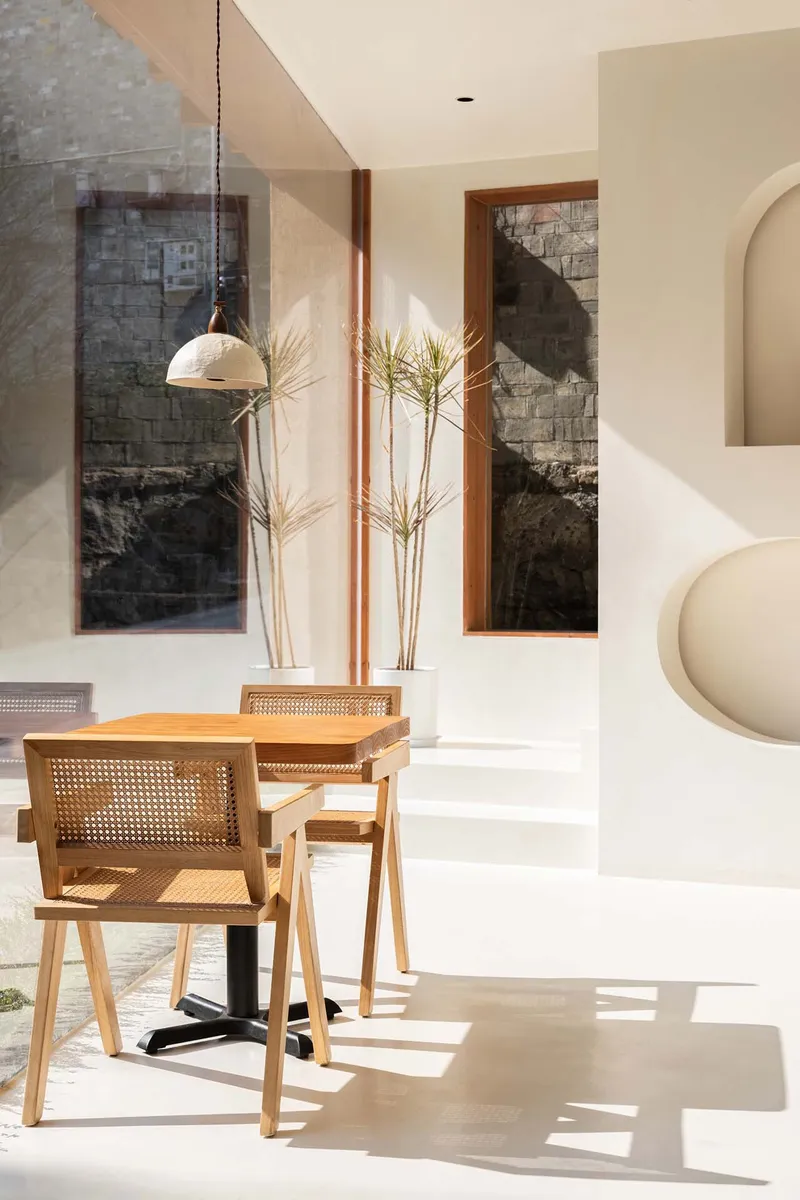 Dining area with cane-back chairs in morning sunlight beside tall windows framing stone courtyard wall
