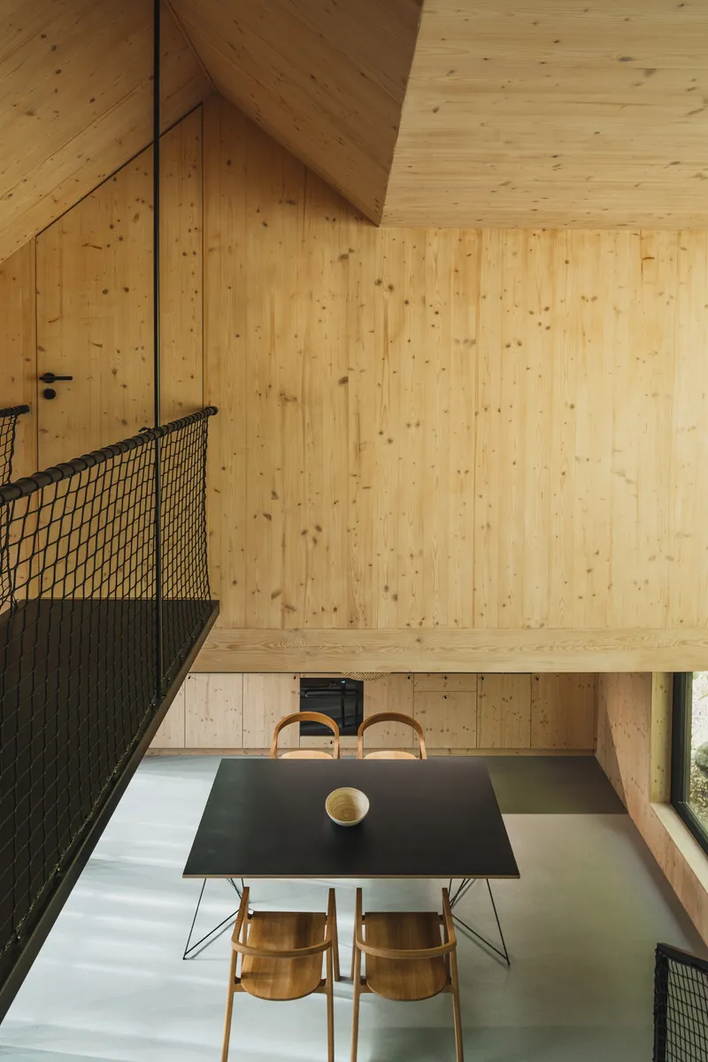 Double-height dining space with a black table below a suspended bridge with mesh balustrade