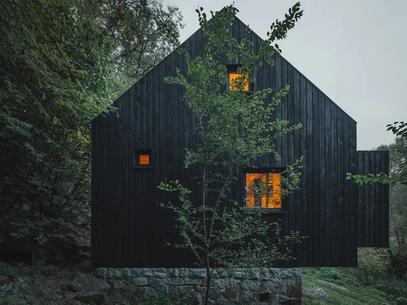 Black vertical timber cladding on a gabled volume with illuminated window openings at dusk