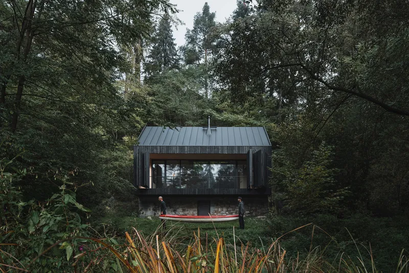 Weathered timber cabin with standing seam metal roof and glazed opening framed by dense forest