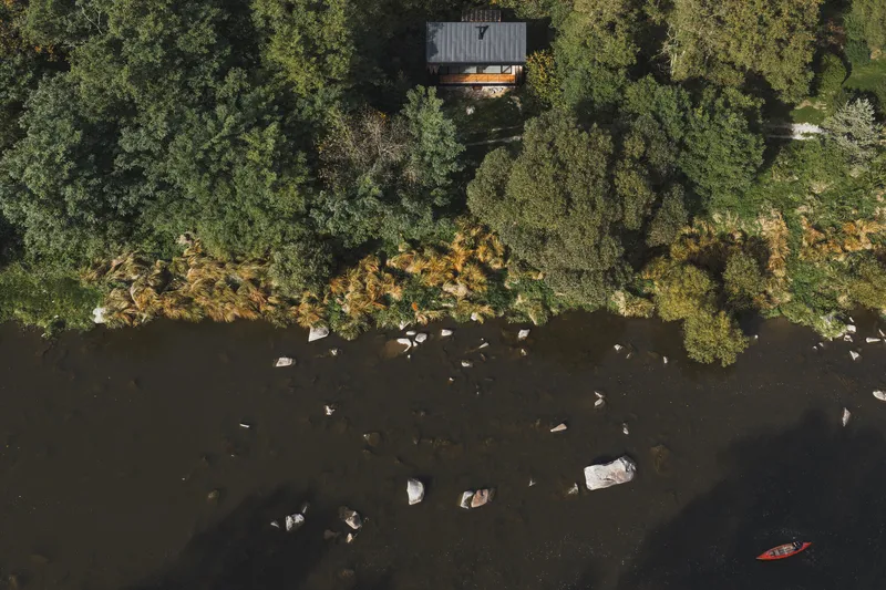 Aerial view of the timber cabin cantilevered above a rocky shoreline with kayaker below