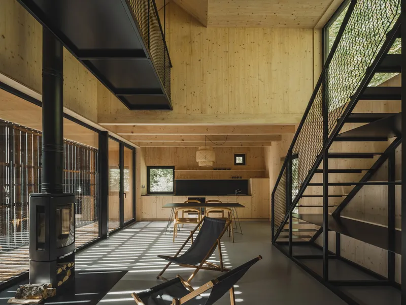 Double-height interior with black steel staircase, wood-burning stove, and plywood-lined walls in striped sunlight