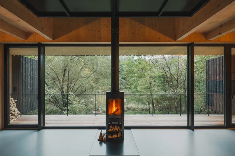 Freestanding wood stove centered before full-height glazing overlooking forest in afternoon light