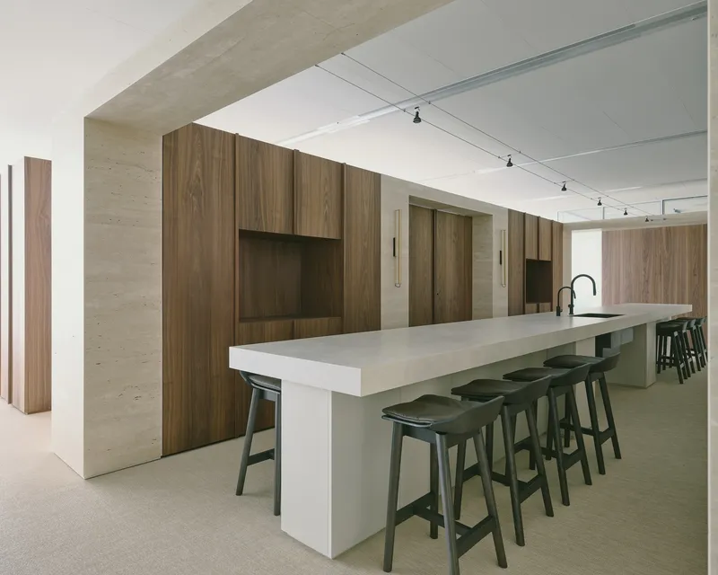 Long kitchen island with pale concrete countertop and black stools beneath track lighting on white ceiling