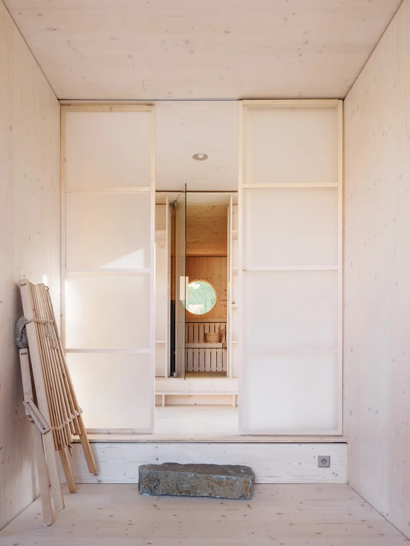 Light plywood anteroom with translucent sliding panels revealing glimpse of sauna space with folded chair