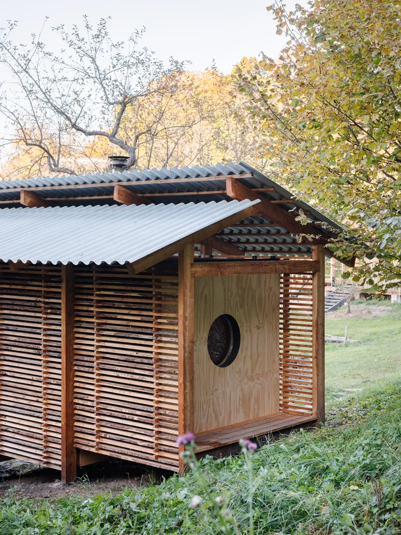 Small structure with corrugated metal roof, plywood walls, and horizontal wood slat screens beneath autumn trees
