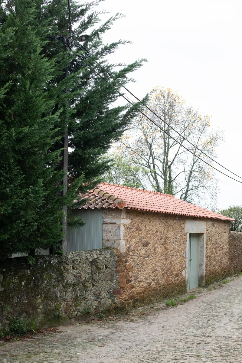 Stone outbuilding with red clay tile roof and sage green corrugated addition beside cobblestone lane