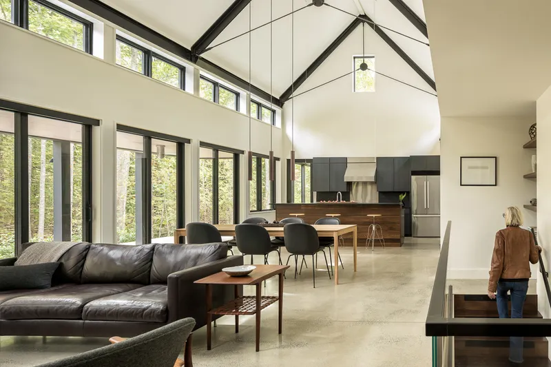 Open living and dining space with exposed timber trusses and floor-to-ceiling glazing overlooking forest