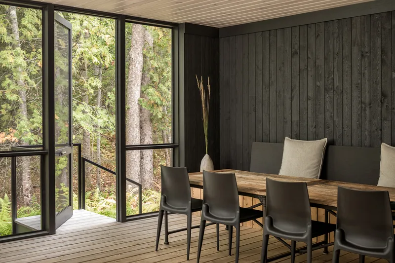 Dining area with charcoal-stained vertical planks and floor-to-ceiling glazing overlooking dense forest