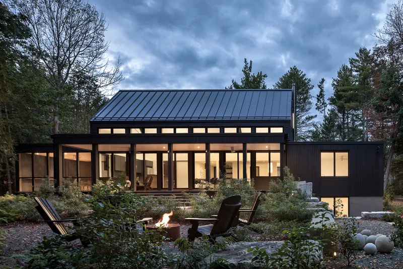 Rear elevation showing standing seam metal roof, glazed openings, and fire pit at twilight