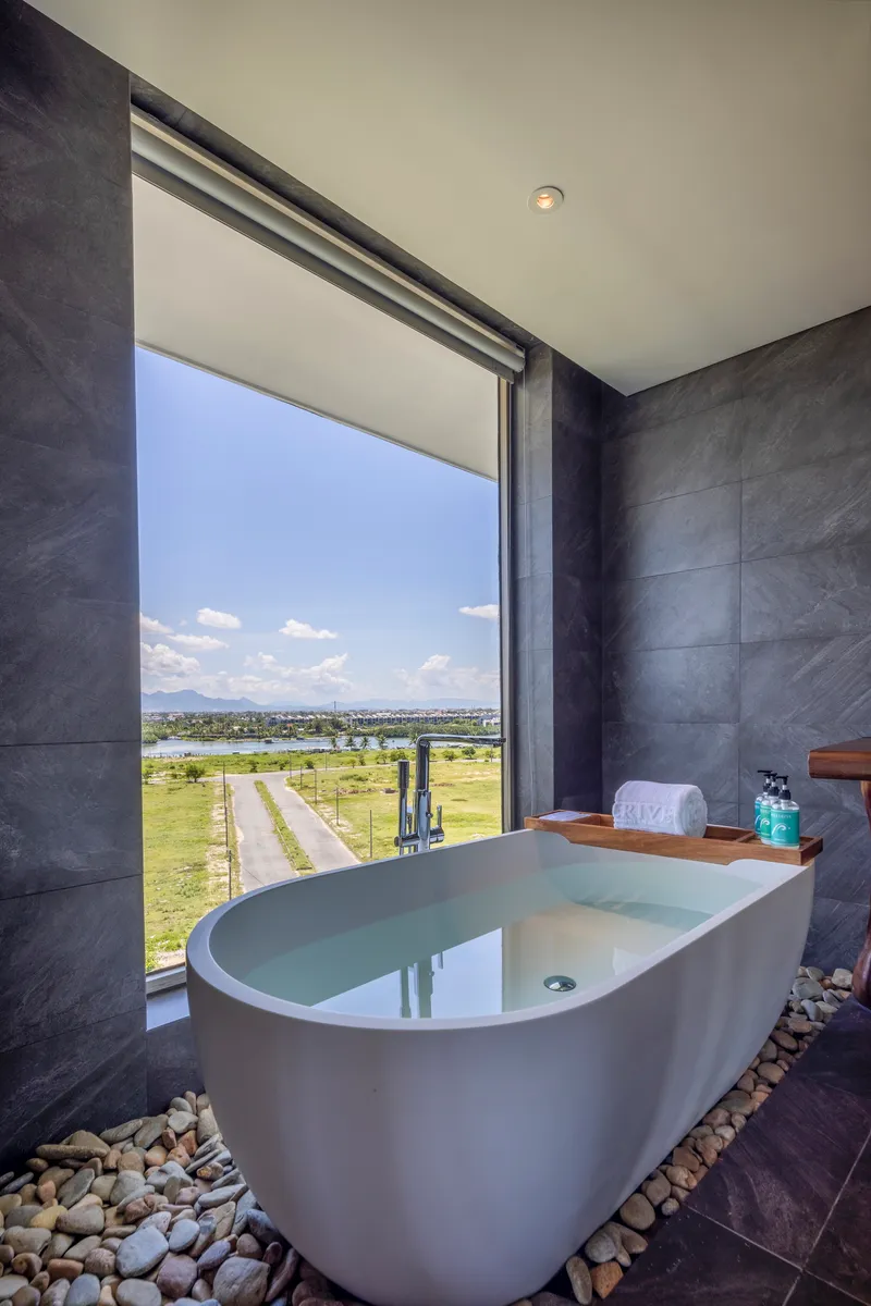 Freestanding bathtub on river stones against dark tile walls with a full-height window framing distant fields