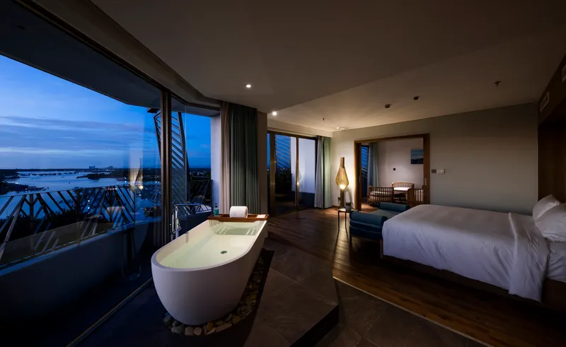 Hotel suite with freestanding bathtub on pebble bed and balcony view over water at dusk