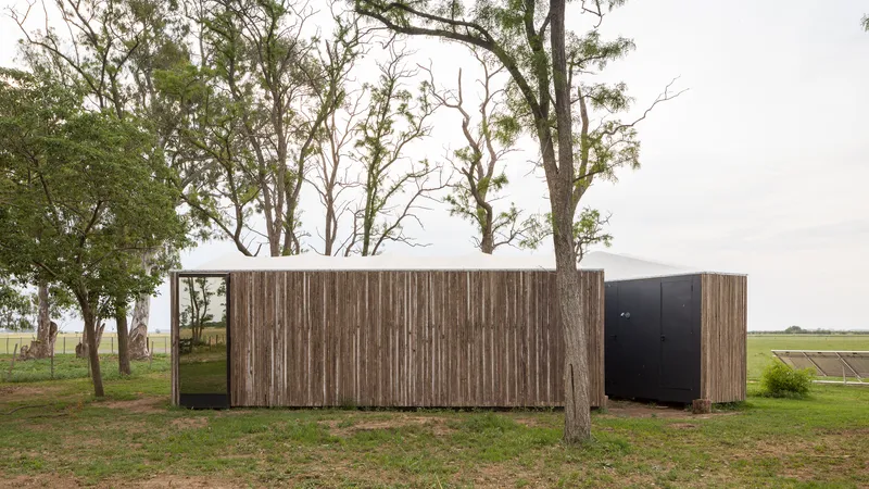 Timber-clad volume with black door nestled among mature trees in an open field