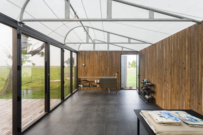 Interior space with exposed steel roof trusses, full-height glazing, vertical timber walls and grey floor tiles