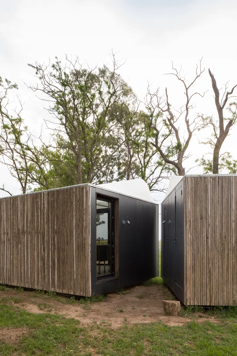 Entry courtyard formed by weathered timber volumes with dark metal pivot door under cloudy sky