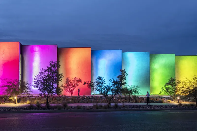 Rainbow-lit concrete wall panels beside a walkway with small trees and a figure at twilight