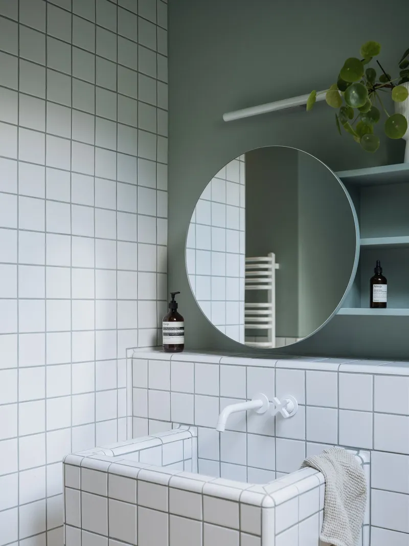 Bathroom corner with white square tile wall, round mirror, and sage green painted alcove with shelving