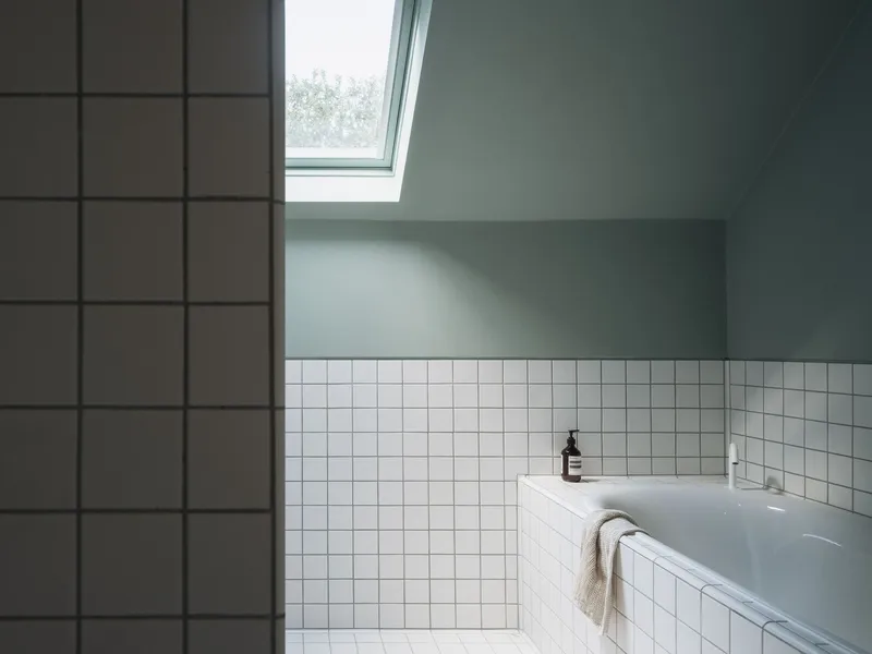 Bathtub corner with white square tiles under a sloped ceiling and angled skylight casting soft daylight