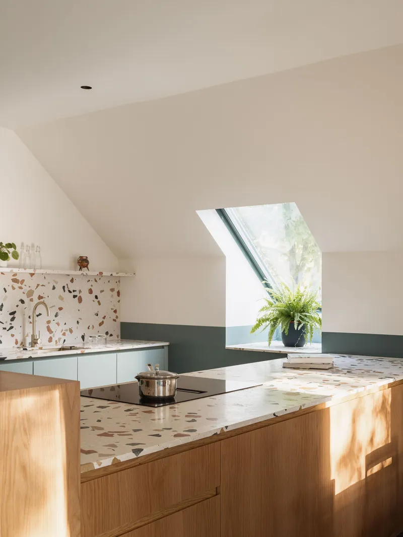 Kitchen workspace with timber cabinets and terrazzo surfaces catching sunlight from the skylight